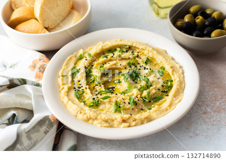 A bowl of creamy hummus topped with olive oil and herbs sits beside fresh bread and a bowl of olives. The light setting showcases a simple, inviting meal A bowl of creamy hummus topped with olive oil and herbs sits beside fresh bread and a bowl of olives. The light setting showcases a simple, inviting meal 132714890