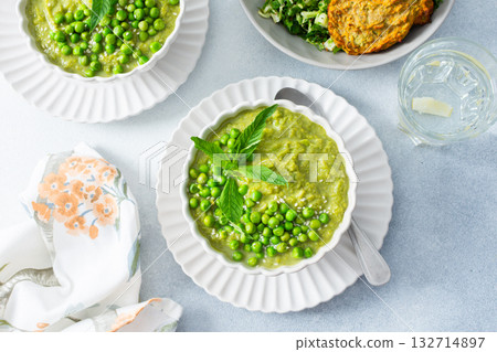 Creamy green pea and broccoli soup garnished with fresh peas is paired with crispy zucchini pancakes and a vibrant salad, creating a healthy and delightful meal 132714897