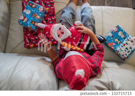 A young boy sits on a sofa holding an inflatable Santa toy, surrounded by Christmas gifts. Festive holiday scene, ideal for Christmas, winter, and seasonal projects. 132715715