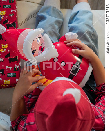 A young boy wearing a Santa hat sits on a sofa, holding an inflatable Santa toy. Cheerful holiday scene, perfect for Christmas, festive, and winter-themed projects. 132715718