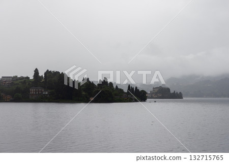 Foggy morning on Lake Orta with misty calm water and distant villas hidden in gentle early haze Foggy morning on Lake Orta with misty calm water and distant villas hidden in gentle early haze 132715765