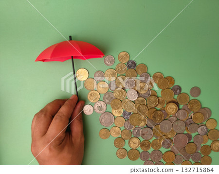 The hand holding a small red umbrella over a pile of coins symbolizes the act of safeguarding one's finances from potential losses or risks. 132715864