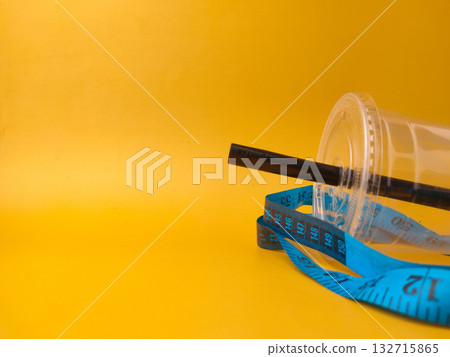 The image shows a clear disposable plastic cup with a black straw, encircled by a blue measuring tape on yellow background. The image shows a clear disposable plastic cup with a black straw, encircled by a blue measuring tape on yellow background. 132715865
