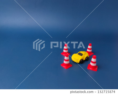 This image shows a small yellow toy car surrounded by four miniature traffic cones on a blue background. This image shows a small yellow toy car surrounded by four miniature traffic cones on a blue background. 132715874