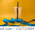 The image shows a clear disposable plastic cup with a black straw, encircled by a blue measuring tape on yellow background. 132715889