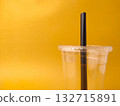 The image shows a clear disposable plastic cup with a lid and a black straw on a yellow background. 132715891