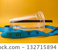 The image shows a clear disposable plastic cup with a black straw, encircled by a blue measuring tape on yellow background. 132715894