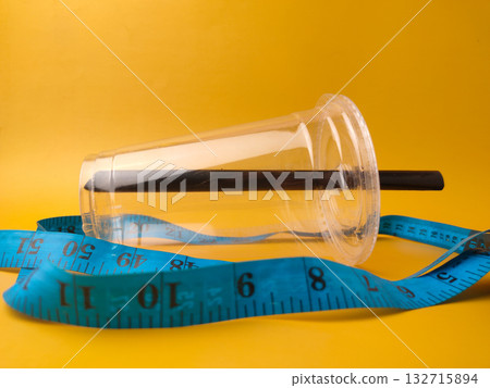 The image shows a clear disposable plastic cup with a black straw, encircled by a blue measuring tape on yellow background. 132715894