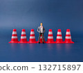 The image shows a small figurine of a man in a business suit standing among a line of red and white traffic cones. 132715897