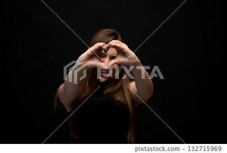 Playful young woman with long brown hair making heart shape with hands on black background 132715969