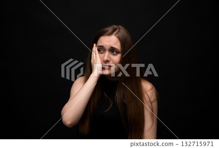 Sad young woman with long brown hair touching face with hand expressing pain on dark background 132715971