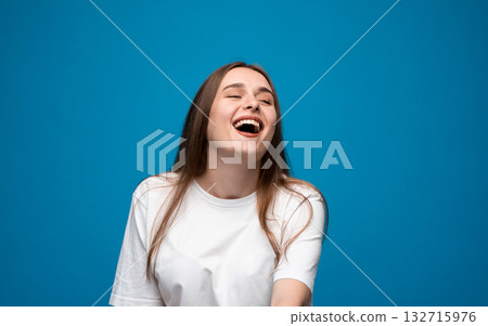 Laughing young woman with long brown hair in white t-shirt expressing joy on blue background Laughing young woman with long brown hair in white t-shirt expressing joy on blue background 132715976