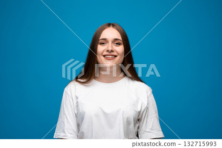 Smiling young woman with long brown hair and blue eyes wearing white t-shirt on blue background 132715993