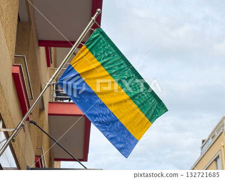 Flag of Gabon waving against the sky. Cultural identity, national pride, and harmony between color, structure, and movement in urban surroundings. 132721685