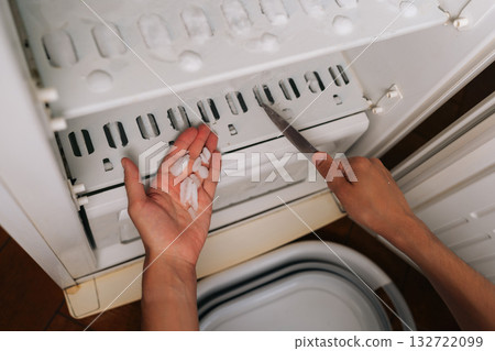 Top view of male hands scraping frost accumulation from refrigerator interior with knife, demonstrating maintenance process for preserving cooling equipment performance. Top view of male hands scraping frost accumulation from refrigerator interior with knife, demonstrating maintenance process for preserving cooling equipment performance. 132722099
