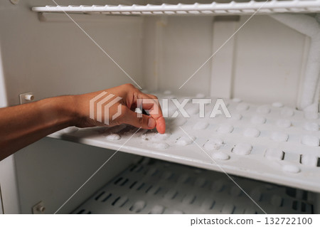 Fingertips pressing against thick frost layer inside refrigerator freezer, revealing heavy ice accumulation requiring urgent defrosting. Concept of need for defrosting due to excessive buildup. 132722100