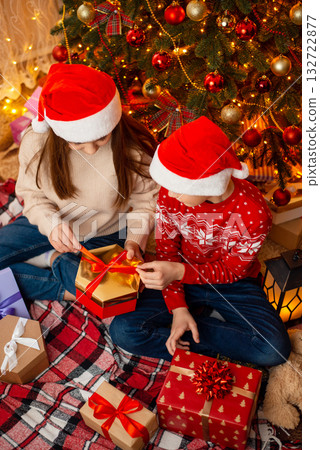 Horizontal photo of two children near the christmas tree, opening presents. Winter holidays, happy family atmosphere, festive interior. 132722877