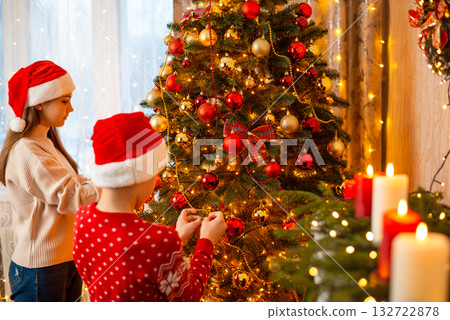 Two siblings children boy and girl in Santa Claus hats and warm sweaters decorate Christmas tree in cozy living room. Celebrating Christmas and New Year with family. Two siblings children boy and girl in Santa Claus hats and warm sweaters decorate Christmas tree in cozy living room. Celebrating Christmas and New Year with family. 132722878