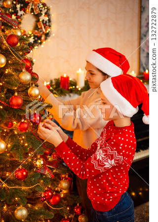 Two happy friends children in Santa Claus hats decorate Christmas tree. Little helpers, spending holidays together, horizontal shot. 132722879