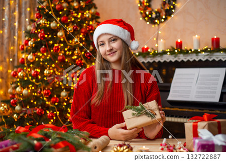 Happy young girl sitting near the table and holding packed and decorated present box. Gorgeous Christmas tree and piano on the background Happy young girl sitting near the table and holding packed and decorated present box. Gorgeous Christmas tree and piano on the background 132722887