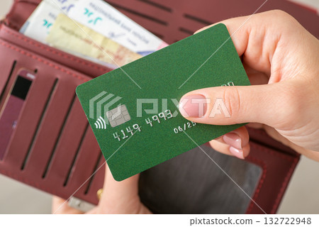 Woman's hands holding a green credit card and burgundy wallet with Ukrainian Hryvnias, close up. Banking, cashless payments, financed, purchases and money concept 132722948