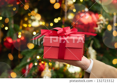 Man hand holding red gift box or present against Christmas tree. Xmas shopping. Boxing Day. Close up. Man hand holding red gift box or present against Christmas tree. Xmas shopping. Boxing Day. Close up. 132723312