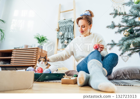 Young Woman in cozy sweater petting cat while sitting on floor cushions and decorating potted Christmas tree in modern Scandi interior home. Eco-friendly cozy winter holidays mood. Selective focus. Young Woman in cozy sweater petting cat while sitting on floor cushions and decorating potted Christmas tree in modern Scandi interior home. Eco-friendly cozy winter holidays mood. Selective focus. 132723313