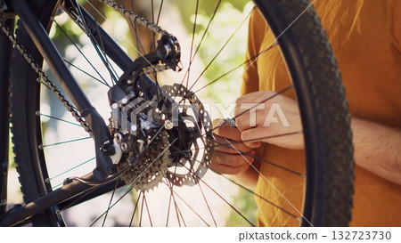 Outdoor close-up of dedicated caucasian man fastening and adjusting the cassette on his bike for annual maintenance. Detailed view of sporty male cyclist examining bicycle wheel in his backyard. 132723730