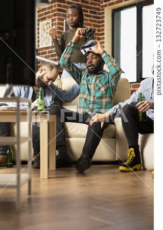 Diverse friends express sadness after video game defeat in cozy apartment. African american man holding VR headset and controller raises them in frustration after losing online gaming tournament. Diverse friends express sadness after video game defeat in cozy apartment. African american man holding VR headset and controller raises them in frustration after losing online gaming tournament. 132723749
