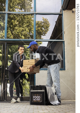 African american man delivering pizza in a large stack of cardboard boxes to a client standing in her doorway. The transaction is part of a routine, fast paced city courier service. 132723763
