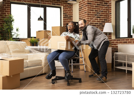 Young multiethnic couple enjoying playful moments in new home, laughing and riding on chair surrounded by cardboard boxes. Happy boyfriend and girlfriend celebrate moving day with fun and excitement. 132723780