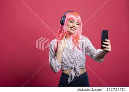 Cheerful woman with pink hair posing for selfie, flashing peace sign and making fun, cheeky face in front of isolated backdrop. Playful young lady with headphones smiling brightly towards cellphone. Cheerful woman with pink hair posing for selfie, flashing peace sign and making fun, cheeky face in front of isolated backdrop. Playful young lady with headphones smiling brightly towards cellphone. 132723781
