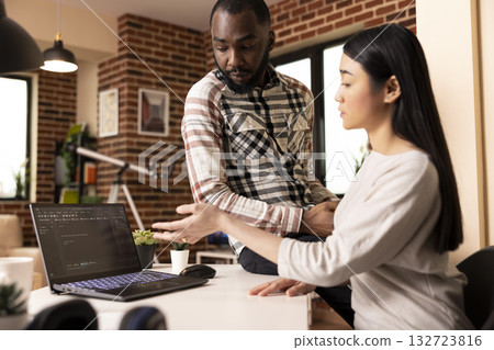Multiethnic couple collaborating in home office, one seated and one standing, reviewing programming content on laptop. Warm lighting, teamwork and diverse partnership in brick wall apartment. 132723816