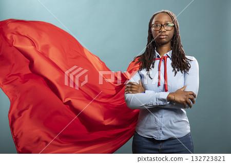 Confident black woman stands in a striking heroic pose, exuding strength and determination. African american lady in a bold red cape and blue shirt, stands with arms crossed in studio. Confident black woman stands in a striking heroic pose, exuding strength and determination. African american lady in a bold red cape and blue shirt, stands with arms crossed in studio. 132723821