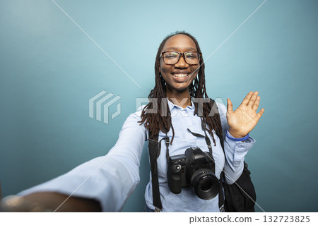 Smiling female photographer makes hello gesture, capturing video content with dslr around her neck. African American woman waving at her online audience, sharing updates of her photography journey. 132723825