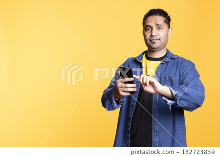 Indian man being calm and smiling interacting with his smartphone, navigating on digital apps and enjoying wireless connectivity. Casual pose against a bright orange studio background. Indian man being calm and smiling interacting with his smartphone, navigating on digital apps and enjoying wireless connectivity. Casual pose against a bright orange studio background. 132723839