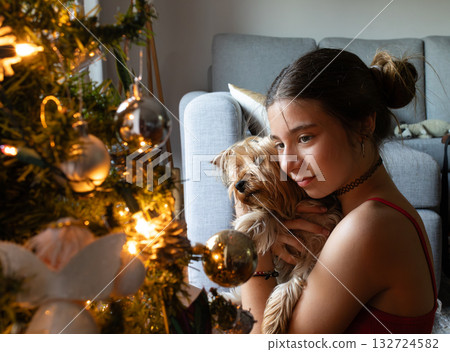 Young woman enjoying festive holiday season at home, cradling cute yawning yorkshire terrier dog next to a decorated christmas tree 132724582