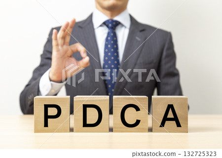 一位商人擺出OK的姿勢,手裡拿著一個寫著「PDCA」的積木。 一位商人擺出OK的姿勢,手裡拿著一個寫著「PDCA」的積木。 132725323
