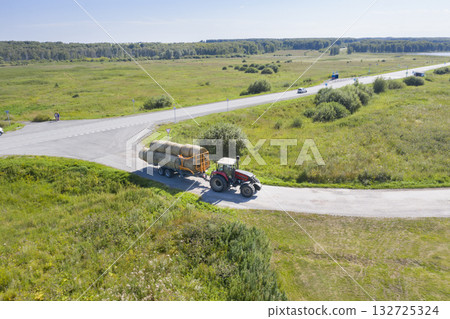 Aerial view of a tractor carrying a huge hay trailer along a country road in summer Aerial view of a tractor carrying a huge hay trailer along a country road in summer 132725324