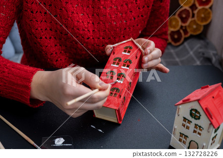 Sustainableminded Christmas cozy atmospheric festive decor in modern scandinavian home handmade decoration. Unrecognizable woman in red sweater painting craft wooden houses. Seasonal activity for kids 132728226