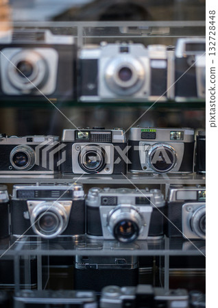 A blurred showcase of vintage cameras behind glass with a reflection of people and the urban landscape. Universal background. Nostalgia, photography, history. High quality photo A blurred showcase of vintage cameras behind glass with a reflection of people and the urban landscape. Universal background. Nostalgia, photography, history. High quality photo 132728448