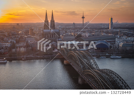 A breathtaking view of the Cologne skyline during sunset featuring the iconic Cologne Cathedral and the city's famous bridge. The warm hues of the evening sky create a stunning scene in Germany. 132728449