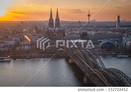 A breathtaking view of the Cologne skyline during sunset featuring the iconic Cologne Cathedral and the city's famous bridge. The warm hues of the evening sky create a stunning scene in Germany. 132728452