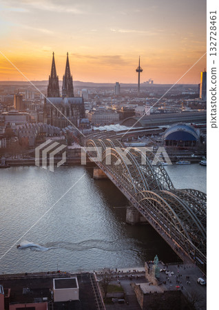 A breathtaking view of the Cologne skyline during sunset featuring the iconic Cologne Cathedral and the city's famous bridge. The warm hues of the evening sky create a stunning scene in Germany. 132728461