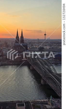 A breathtaking view of the Cologne skyline during sunset featuring the iconic Cologne Cathedral and the city's famous bridge. The warm hues of the evening sky create a stunning scene in Germany. A breathtaking view of the Cologne skyline during sunset featuring the iconic Cologne Cathedral and the city's famous bridge. The warm hues of the evening sky create a stunning scene in Germany. 132728494