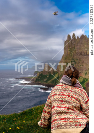 Girl sitting and watching Drangarnir cliff with Tindholmur islands in the background during early morning hike in summer at Faroese coastline Faroe Island, Denmark, Europe. High quality photo 132728585