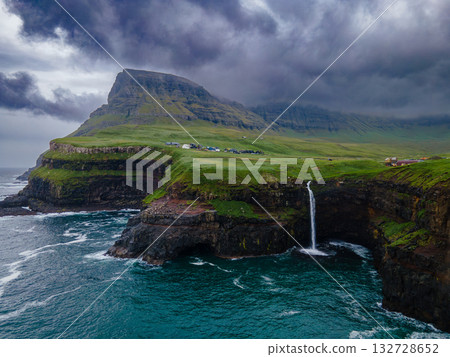 Dramatic Mulafossur waterfall plunging into the ocean with Gasadalur village and cliffs in the background. Dramatic Mulafossur waterfall plunging into the ocean with Gasadalur village and cliffs in the background. 132728652