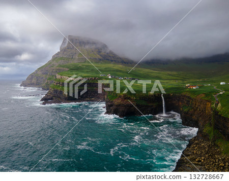 Dramatic Mulafossur waterfall plunging into the ocean with Gasadalur village and cliffs in the background. Dramatic Mulafossur waterfall plunging into the ocean with Gasadalur village and cliffs in the background. 132728667