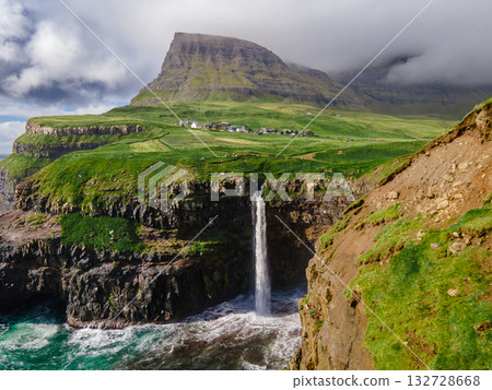 Dramatic Mulafossur waterfall plunging into the ocean with Gasadalur village and cliffs in the background. 132728668