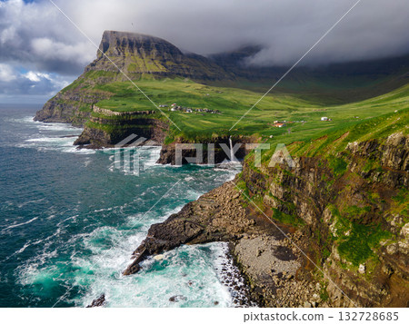 Dramatic Mulafossur waterfall plunging into the ocean with Gasadalur village and cliffs in the background. Dramatic Mulafossur waterfall plunging into the ocean with Gasadalur village and cliffs in the background. 132728685